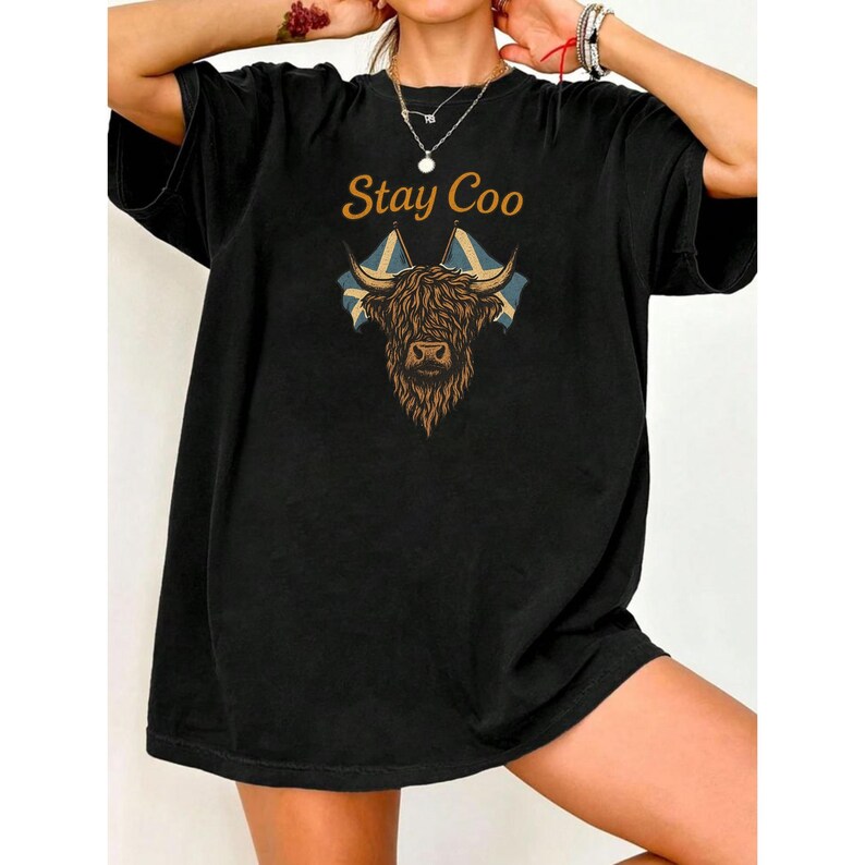 K&ouml;nnte beinhalten: Schwarzes T-Shirt mit dem Text "Stay Coo" &uuml;ber einer Grafik einer Highland-Kuh mit schottischen Flaggen. Die Kuh ist braun und hellbraun, die Flaggen blau und wei&szlig;.