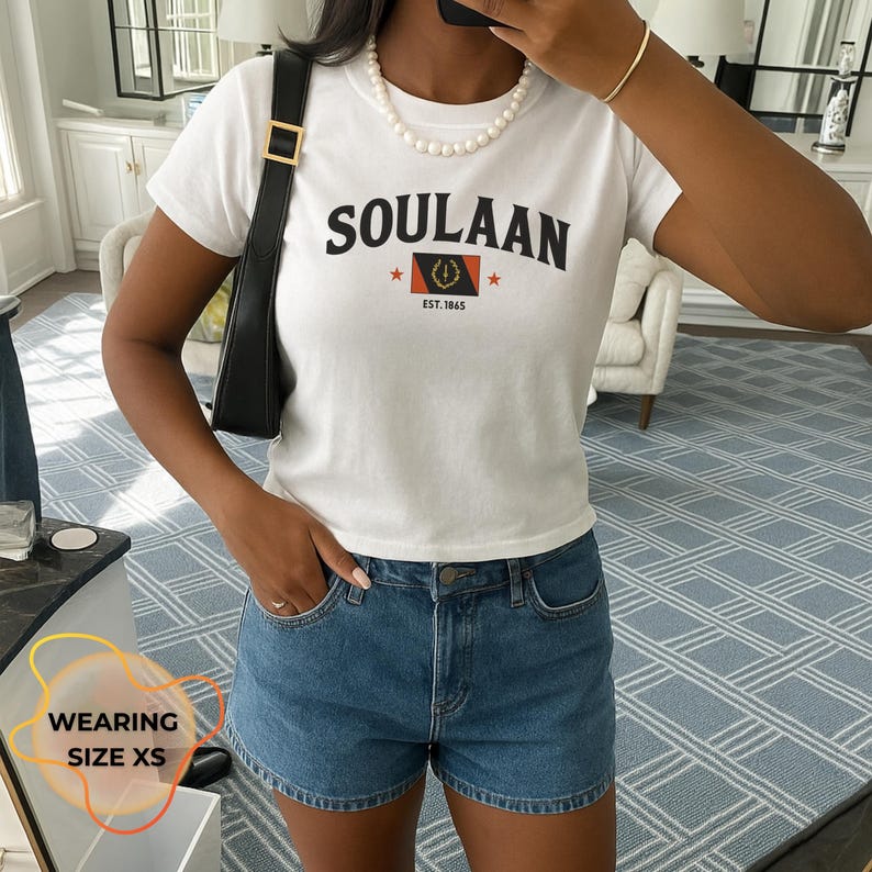 May include: White t-shirt with the word "SOULAAN" in black block letters. Below the word is a graphic with a red star on each side and a flag design. The shirt is paired with blue denim shorts.