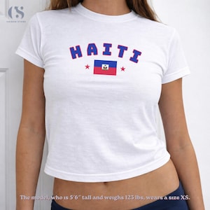 May include: White t-shirt with the word "HAITI" in blue letters and a red outline. The shirt also features the Haitian flag and red stars. A casual, crew neck tee.