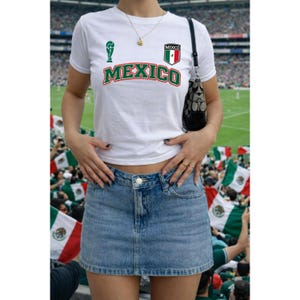 Op de afbeelding: Een wit T-shirt met korte mouwen met het woord MEXICO in groene en rode letters, een groen trofee-icoon en een klein Mexicaans vlag-embleem op de borst, gecombineerd met een blauwe spijkerrok en een zwarte schoudertas met een patroon.