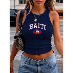 Puede incluir: Una camiseta de tirantes acanalada de color azul oscuro con la palabra HAITI en letras mayúsculas blancas arqueadas sobre un pequeño gráfico ovalado de la bandera de Haití. La prenda se combina con vaqueros azules y un bolso negro estampado.