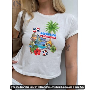 Op de afbeelding: Wit t-shirt met een kleurrijke afbeelding van de Dominicaanse Republiek, inclusief vlaggen, een palmboom en de tekst "República Dominicana". Het ontwerp bevat een kaartomtrek en nationale symbolen.