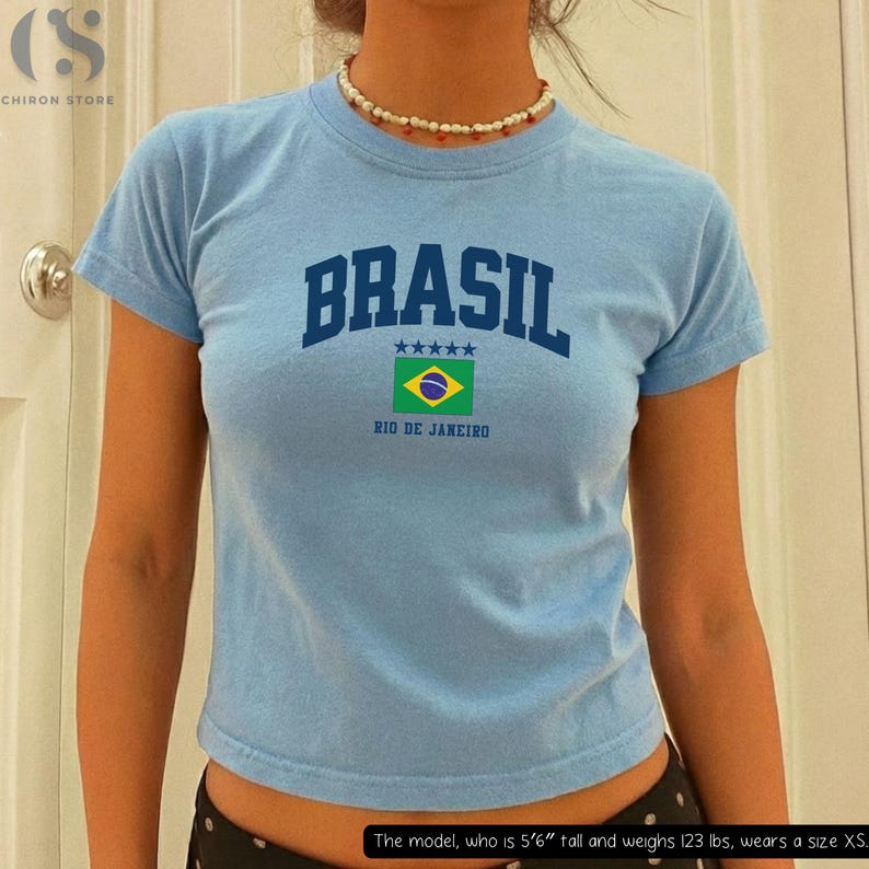May include: Light blue t-shirt with the word "BRASIL" in navy blue block letters, a Brazilian flag graphic, and the text "RIO DE JANEIRO." The shirt has short sleeves and a crew neck.