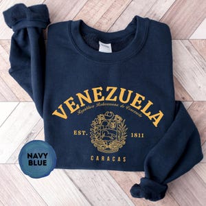 May include: Navy blue sweatshirt with gold lettering and a crest design. The word "VENEZUELA" arches across the top, with "Rep&uacute;blica Bolivariana de Venezuela" below. The crest includes a horse, trees, and the text "EST. 1811 CARACAS".