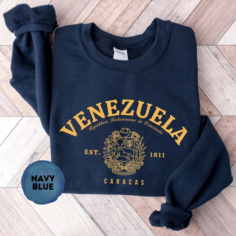 May include: Navy blue sweatshirt with gold lettering and a crest design. The word "VENEZUELA" arches across the top, with "Rep&uacute;blica Bolivariana de Venezuela" below. The crest includes a horse, trees, and the text "EST. 1811 CARACAS".