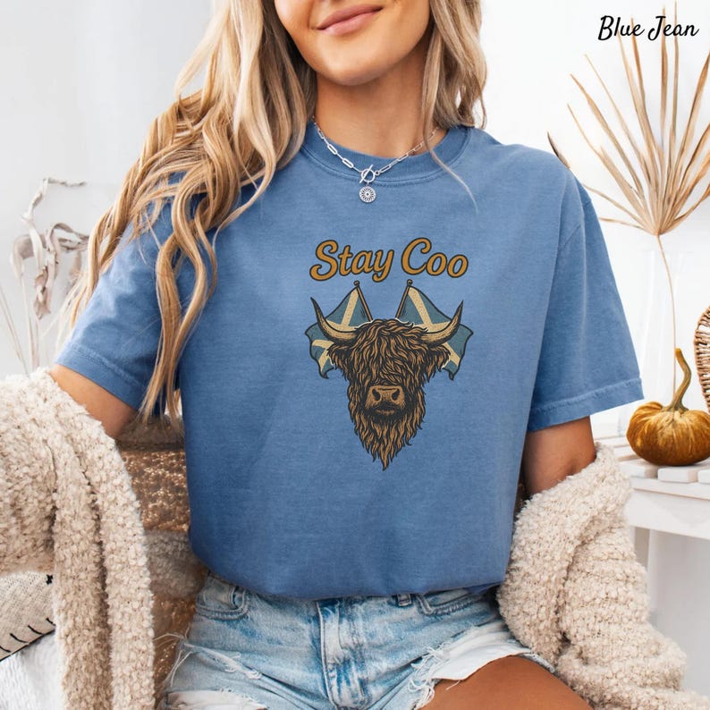 K&ouml;nnte beinhalten: Blaues Jeans-T-Shirt mit einer Illustration einer Highland-Kuh, schottischen Flaggen und dem Text "Stay Coo". Das Design verwendet Braun-, Beige- und Blaut&ouml;ne. Das Shirt hat kurze &Auml;rmel und einen Rundhalsausschnitt.