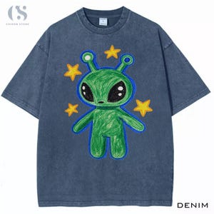 Może przedstawiać: Ciemnoniebieska koszulka typu t-shirt z efektem sprania, z centralną grafiką przedstawiającą zielonego kosmitę z dużymi czarnymi oczami i czułkami. Kosmita jest otoczony czterema żółtymi gwiazdami. Wzór ma teksturę przypominającą rysunek ręczny.