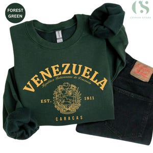 May include: Forest green sweatshirt with "VENEZUELA" in gold lettering. The design includes the text "Rep&uacute;blica Bolivariana de Venezuela", "EST. 1811", and "CARACAS". A pair of dark wash jeans is next to the sweatshirt.
