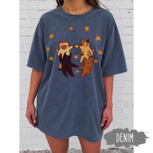 May include: A denim blue short-sleeve t-shirt featuring a graphic print of four cats in various colors holding paws in a circle, surrounded by yellow star illustrations. The word DENIM is printed in white on a gray brushstroke background.