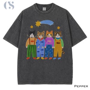 May include: A charcoal grey acid-wash t-shirt featuring a graphic of four cats wearing colourful overalls and sweaters. The cats are positioned beneath a yellow shooting star and two smaller stars. The word PEPPER is printed in the bottom right corner.
