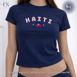 May include: Navy blue crop top with the word "HAITI" in white and red lettering, along with the Haitian flag. The shirt has a crew neck and short sleeves. The model is 5'6" tall and weighs 123 lbs, wearing a size XS.