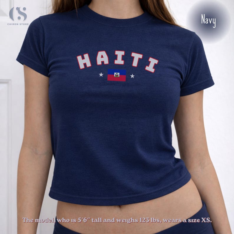 May include: Navy blue crop top with the word "HAITI" in white and red lettering, along with the Haitian flag. The shirt has a crew neck and short sleeves. The model is 5'6" tall and weighs 123 lbs, wearing a size XS.