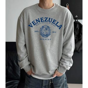 May include: A gray sweatshirt with the word "VENEZUELA" in blue arched across the chest. Below the word is a crest with the words "EST. 1811" and "CARACAS". The sweatshirt is a crew neck style.