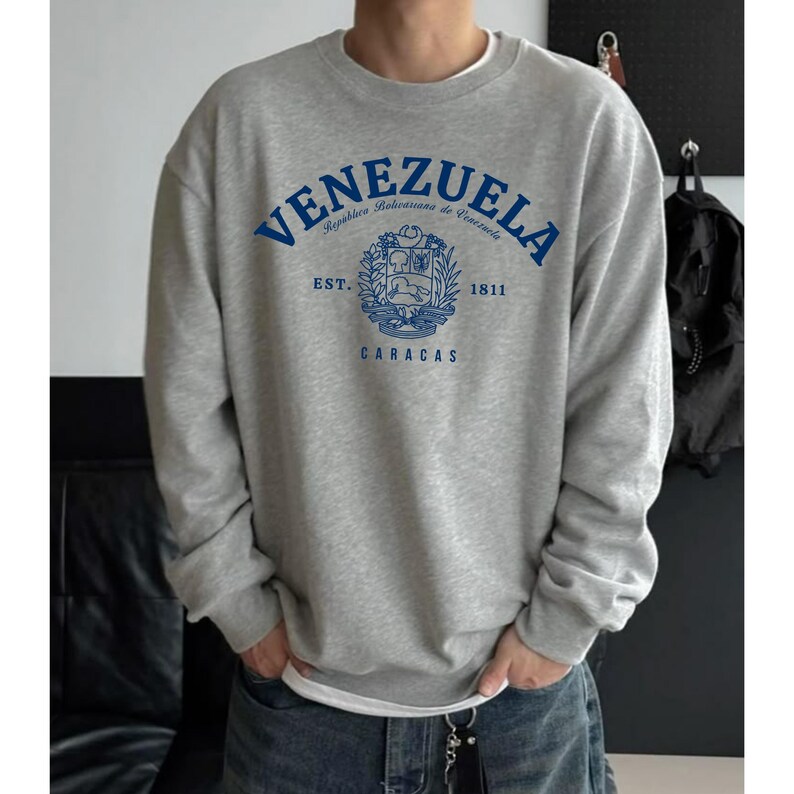 May include: A gray sweatshirt with the word "VENEZUELA" in blue arched across the chest. Below the word is a crest with the words "EST. 1811" and "CARACAS". The sweatshirt is a crew neck style.