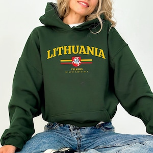 Puede incluir: Una sudadera con capucha verde oscuro con la palabra "LITHUANIA" en letras amarillas. Debajo, un gráfico del escudo de armas lituano y el nombre de la ciudad "VILNIUS". La sudadera tiene una capucha con cordón y un bolsillo delantero.