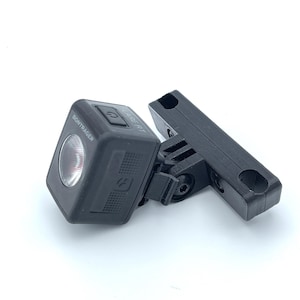 Puede incluir: Una luz trasera de bicicleta Bontrager Flare RT negra con un soporte de montaje. La luz tiene forma cuadrada con esquinas redondeadas y una lente transparente. El soporte también es negro y tiene dos agujeros.