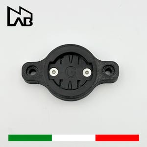 Könnte beinhalten: Schwarze Fahrradcomputerhalterung mit kreisförmiger Mitte und zwei Schraubenlöchern. Die Mitte zeigt den Buchstaben "G" und zwei silberne Schrauben. Die Halterung hat eine rechteckige Form mit abgerundeten Kanten. Das Logo "M AB" befindet sich oben links.