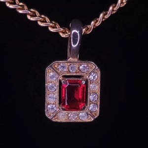 Puede incluir: Un collar de cadena dorada con un colgante que presenta una piedra preciosa roja rodeada de diamantes de imitación transparentes.