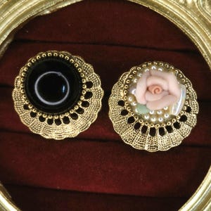 Puede incluir: Dos botones decorativos de color dorado con diseños intrincados. Un botón presenta una piedra negra en cabujón, mientras que el otro tiene una rosa rosa con detalles de perlas.