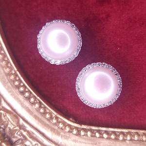 May include: Two round, pearl-like earrings with a halo of small, sparkling crystals. The earrings are set against a deep red velvet background, with a gold-colored ornate frame in the foreground. The earrings have a classic, elegant design.