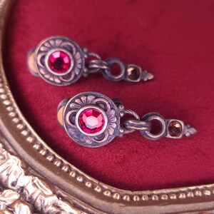 Könnte beinhalten: Ein Paar antike silberfarbene Ohrringe mit einem floralen Design um einen zentralen, runden, roten Edelstein. Die Ohrringe sind durch eine Kette verbunden und haben am Ende einen kleinen, bernsteinfarbenen Stein. Sie werden auf rotem Samt präsentiert.