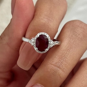 May include: A silver ring featuring a large oval ruby set within a halo of smaller diamonds. The ring has two smaller diamonds on either side of the ruby.