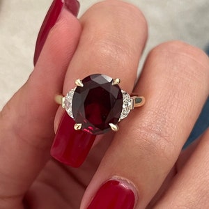 Puede incluir: Un anillo de oro con una gran piedra roja ovalada rodeada de diamantes más pequeños.