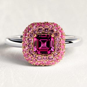 Peut inclure: Bague en argent ornée d'une pierre rose foncée carrée au centre, entourée d'un halo de petites pierres roses. La bague présente un sertissage doré autour de la pierre centrale et un anneau argenté poli. Design élégant et attrayant.