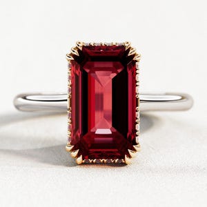 以下が含まれることがあります： 長方形カットの深紅色の宝石リング。 宝石は、小さなアクセントストーンが付いた金色のベゼルにセットされています。 バンドは銀色です。 リングは白い表面に表示されています。