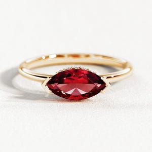 Puede incluir: Un anillo de oro con una gema roja oscura con corte marquesa. La gema es el punto focal, engastada en una delicada banda de oro. El anillo se exhibe sobre una superficie blanca, resaltando su elegante diseño y su color vibrante.
