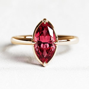 Puede incluir: Un anillo de oro con una gema roja oscura de talla marquesa. La gema está engastada en un delicado bisel de oro, con una banda simple de oro pulido. El anillo se muestra sobre una superficie blanca, resaltando su elegante diseño.