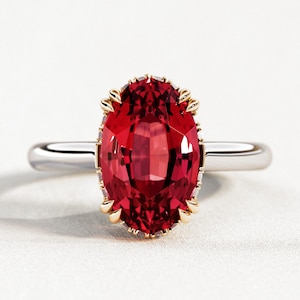 May include: A ruby-red oval gemstone ring with a gold-colored setting and a silver band. The gemstone is the focal point, showcasing a vibrant, faceted surface. The ring is set against a white background.