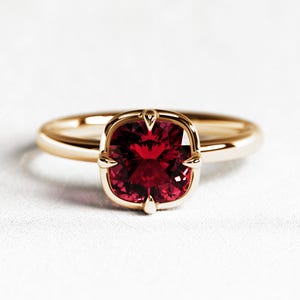 Puede incluir: Un anillo de oro con una piedra preciosa roja oscura de talla cuadrada. La banda y el engaste del anillo son de color oro pulido, con pequeñas garras decorativas que aseguran la piedra. El anillo está sobre un fondo blanco.