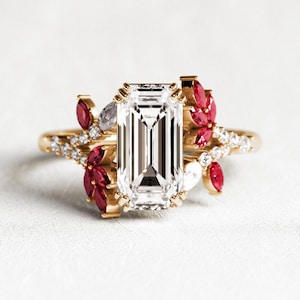 Puede incluir: Un anillo de oro con un diamante central de talla esmeralda, rodeado de piedras preciosas rojas y blancas. La banda presenta pequeños diamantes. El anillo se presenta sobre un fondo blanco, mostrando su intrincado diseño y brillo.