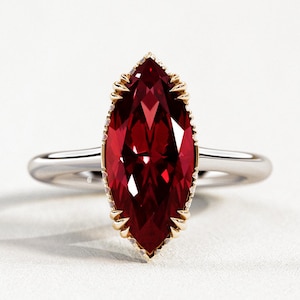 May include: A marquise-cut ruby ring with a gold bezel and a silver band. The ruby is a deep red colour and is faceted. The ring is set against a white background.