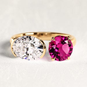 May include: A gold bypass ring featuring two prominent gemstones: a clear, oval-cut stone on the left and a vibrant, round-cut magenta stone on the right. The band is polished gold, and both stones are held in place by secure metal settings.