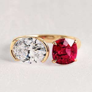 Può includere: Un anello bypass in oro con una pietra preziosa trasparente taglio ovale incastonata a lunetta e una pietra rosso intenso taglio cuscino fissata con quattro griffe. La fascia è in oro lucido e presenta un design aperto con le due pietre.