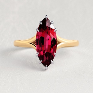May include: A marquise-cut deep red gemstone ring set in a polished yellow gold band. The stone is secured by silver-toned prongs that extend from the center setting, creating a sharp, elongated silhouette against the smooth, solid gold shank.