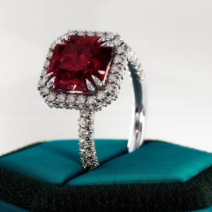 Pode incluir: Anel com uma pedra vermelha rubi de corte quadrado, rodeada por um halo de pequenos diamantes. A banda do anel também é cravejada com diamantes. Exibido sobre uma superfície de veludo verde-azulado.