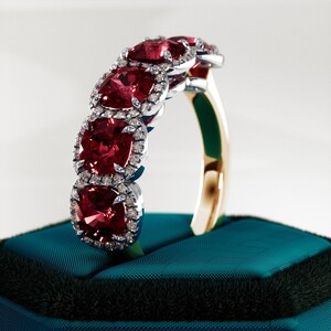 Può includere: Un anello con cinque pietre preziose rosse quadrate, ciascuna circondata da un alone di piccole pietre chiare. La fascia è dorata, con un'incastonatura argentata per le pietre. L'anello è esposto su una superficie di velluto verde acqua.