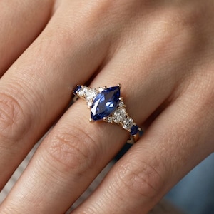 Op de afbeelding: Een marquise-geslepen saffieren ring met een gouden band. De ring heeft een grote, diepblauwe saffieren middensteen, omringd door kleinere witte en blauwe accentstenen. De ring zit aan een hand met gepolijste nagels.