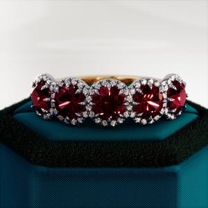 Può includere: Un anello con cinque pietre preziose rosse circondate da pietre preziose bianche più piccole. L'anello è incastonato su una fascia dorata e riposa su una scatola di velluto verde scuro.