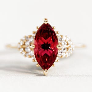 May include: A gold ring featuring a large, marquise-cut, deep red gemstone as the centerpiece. The gemstone is surrounded by smaller, clear gemstones and set on a delicate gold band. The ring has a classic, elegant design.