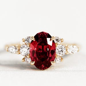 Könnte beinhalten: Ein goldener Ring mit einem großen, oval geschliffenen, tiefroten Edelstein als Mittelpunkt. Der Ring ist mit kleineren, runden, klaren Edelsteinen verziert. Die Fassung ist goldfarben, vor weißem Hintergrund.