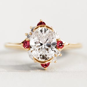 Puede incluir: Un anillo de compromiso de oro con una piedra central de diamante de talla ovalada. El anillo está adornado con piedras preciosas rojas más pequeñas y diamantes blancos, dispuestos en halo. La banda es de oro pulido simple.