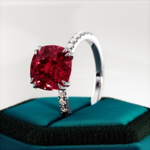 May include: A ring with a large, deep red gemstone set in a white metal band. The band is accented with small, clear gemstones.