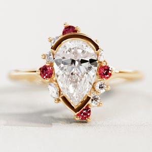 May include: A gold engagement ring featuring a pear-shaped diamond center stone. The ring is accented with red and white gemstones, set in a halo design around the main stone. The band is a delicate gold color.