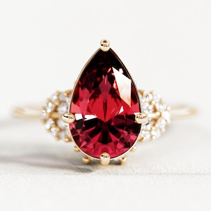 以下が含まれることがあります： 大きな洋梨型の深い赤色の宝石をあしらったゴールドリング。 宝石は金のベゼルにセットされ、小さな輝く白い宝石に囲まれています。 リングのバンドも金です。