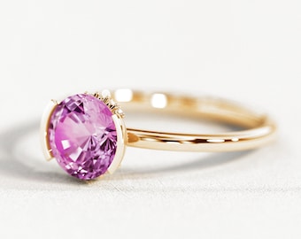 May include: A gold ring featuring a large, faceted, oval-cut pink gemstone. The gemstone is set in a gold bezel setting, and the band is a simple, polished gold. The ring is displayed on a white surface.