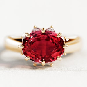 May include: A gold ring featuring a large, oval-cut, deep red gemstone. The gemstone is surrounded by smaller, clear stones and set in a gold band. The ring is displayed on a white surface.